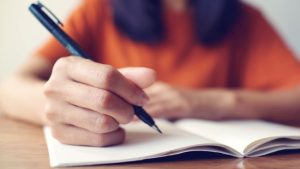 A woman is leaning on desk writing a letter with a pen in a notebook.