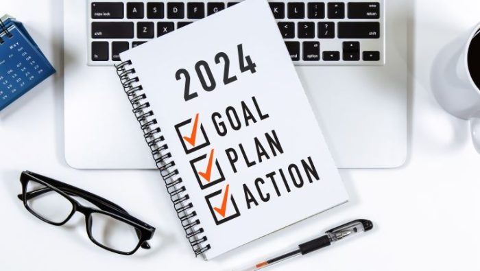 How To Set Achievable Business Goals In 2024 - a open notebook with 2024 goals written in it resting open on a work desk with a laptop and pens.